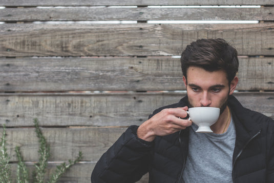 Ein Mann sitzt an einem hohen Holzzaun und trinkt einen Tee aus einer weißen Tasse.