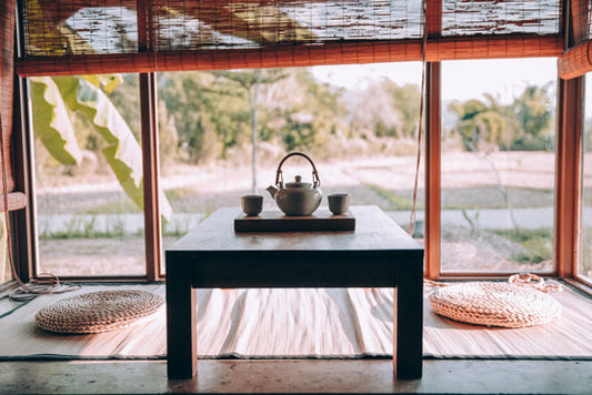 Auf einem Tisch in einem Wintergarten steht eine Teekanne mit 2 Bechern auf einer Holzplatte. Im Wintergarten liegen auch zwei gehäkelte Kissen. Die Sicht nach draussen ist unscharf, man kann aber erkennen, das es sich in einer warmen Gegend abspielt.